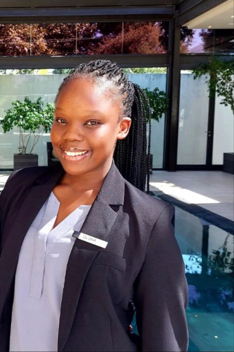 Smiling young woman wearing a black blazer and name tag, standing in a modern indoor setting. - International Hotel School