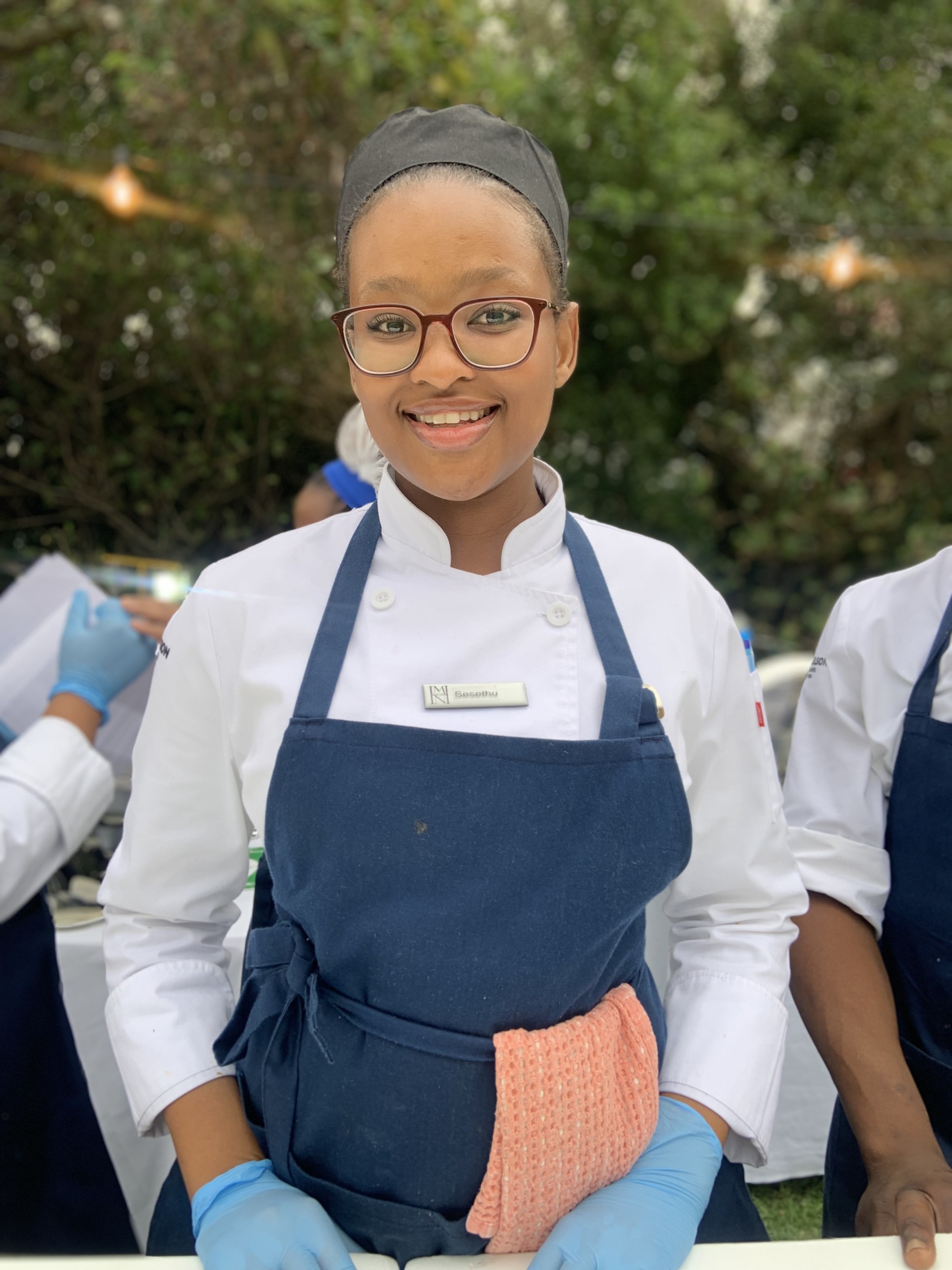 Culinary student wearing a chef uniform and glasses, smiling while preparing food. - International Hotel School