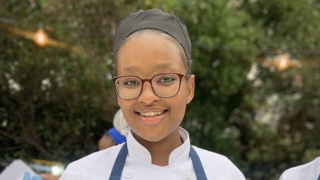 Smiling young female chef wearing glasses and a chef's uniform with a black hat. - International Hotel School