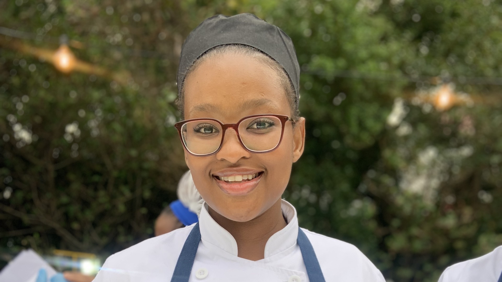 Smiling young female chef wearing glasses and a chef's uniform with a black hat. - International Hotel School