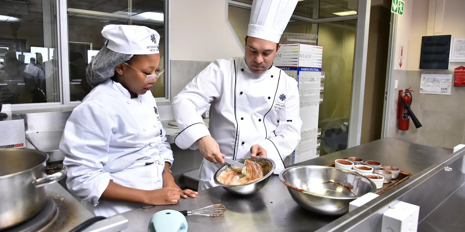 Chef instructing a student in a professional kitchen during a cooking class. - International Hotel School