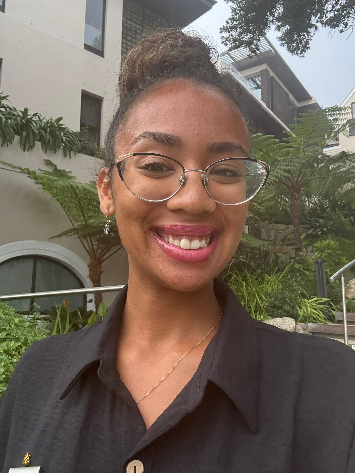 Young woman wearing glasses and a black shirt, smiling outdoors with greenery in the background. - International Hotel School