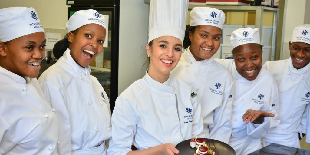 Group of culinary students wearing chef uniforms and smiling while presenting a dish at International Hotel School. - International Hotel School